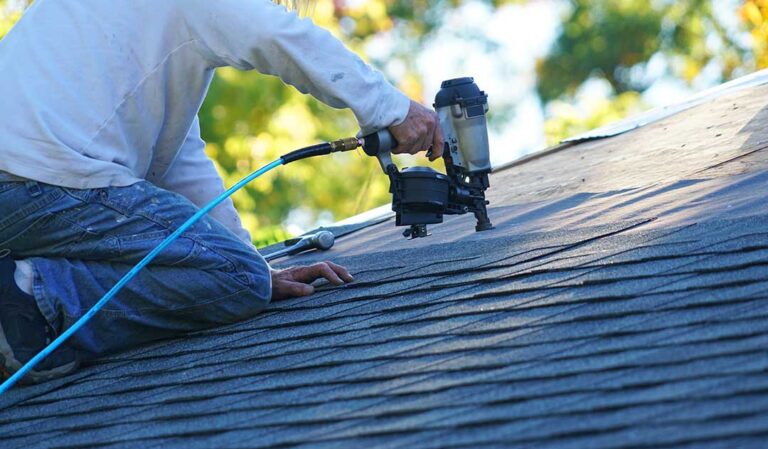 A homeowner applying sealant around a vent pipe on an asphalt shingle roof with gloved hands.