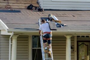 Workers install roofing while attic insulation and ventilation improvements are visible beneath the roof deck.