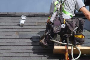 Person clearing leaves from a roof gutter beside shingles, downspout, ladder, and damp autumn ground.
